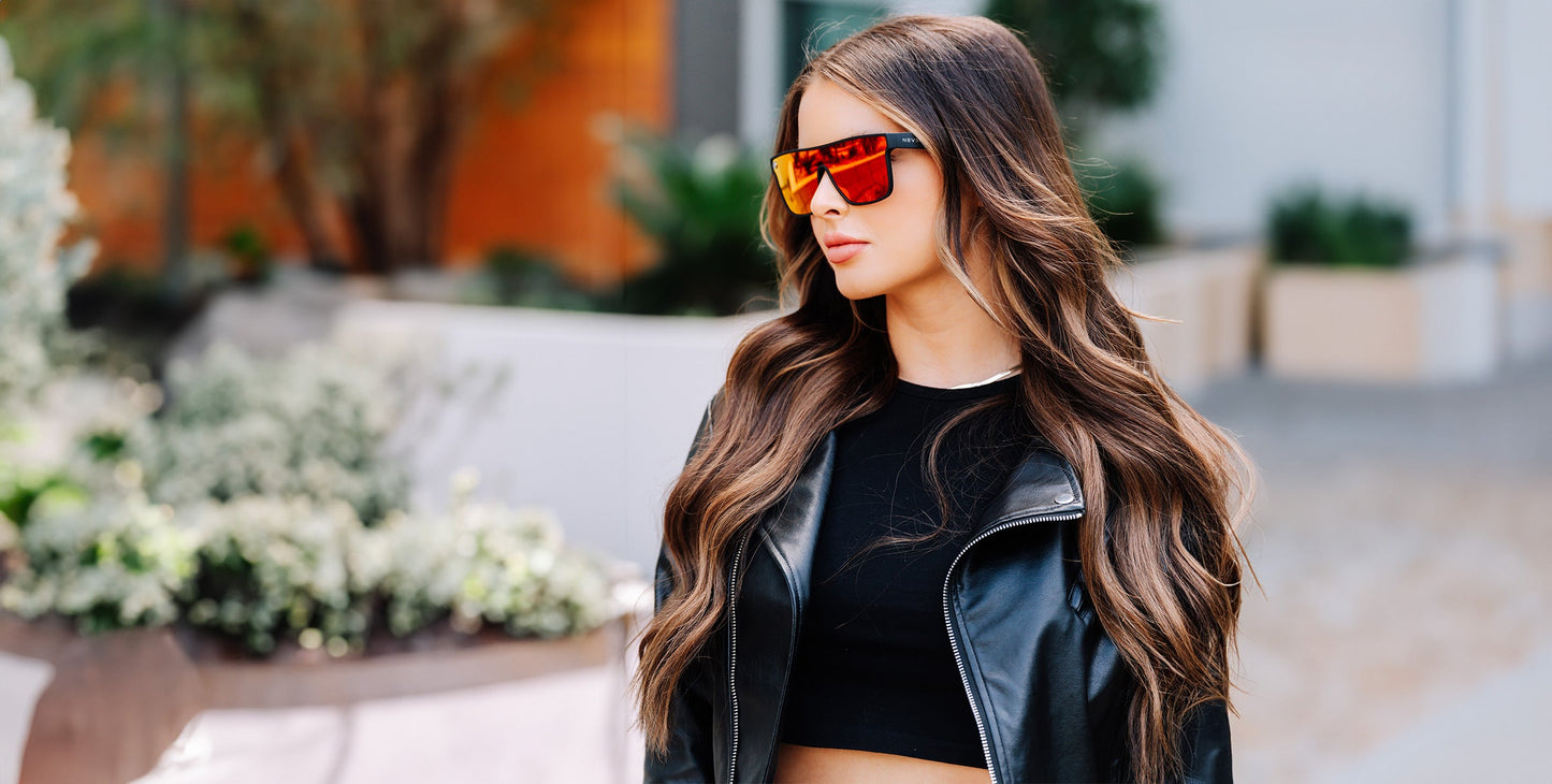 A young woman with long wavy brown hair confidently stands outdoors near plants and modern buildings, wearing Neven Eyewear's Zero Cool cyberpunk sunglasses, a black crop top, and a black leather jacket.