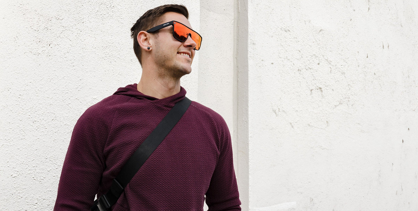 A man in a maroon textured hoodie and black crossbody bag stands against a white wall, smiling to the side. He wears short brown hair, a small earring, and rocks Neven Eyewear's Zero Cool large reflective orange sunglasses.