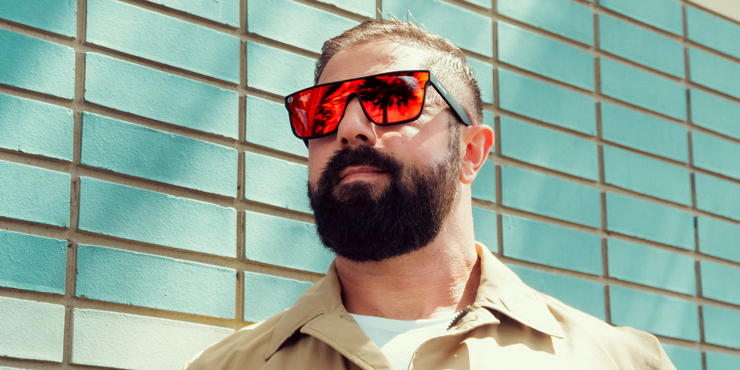 A man with a full beard and short hair wears Neven Eyewear's Zero Cool XL oversized red sunglasses with UV400 protection and a beige collared shirt. He stands before a teal brick wall, palm trees reflecting in his shades.