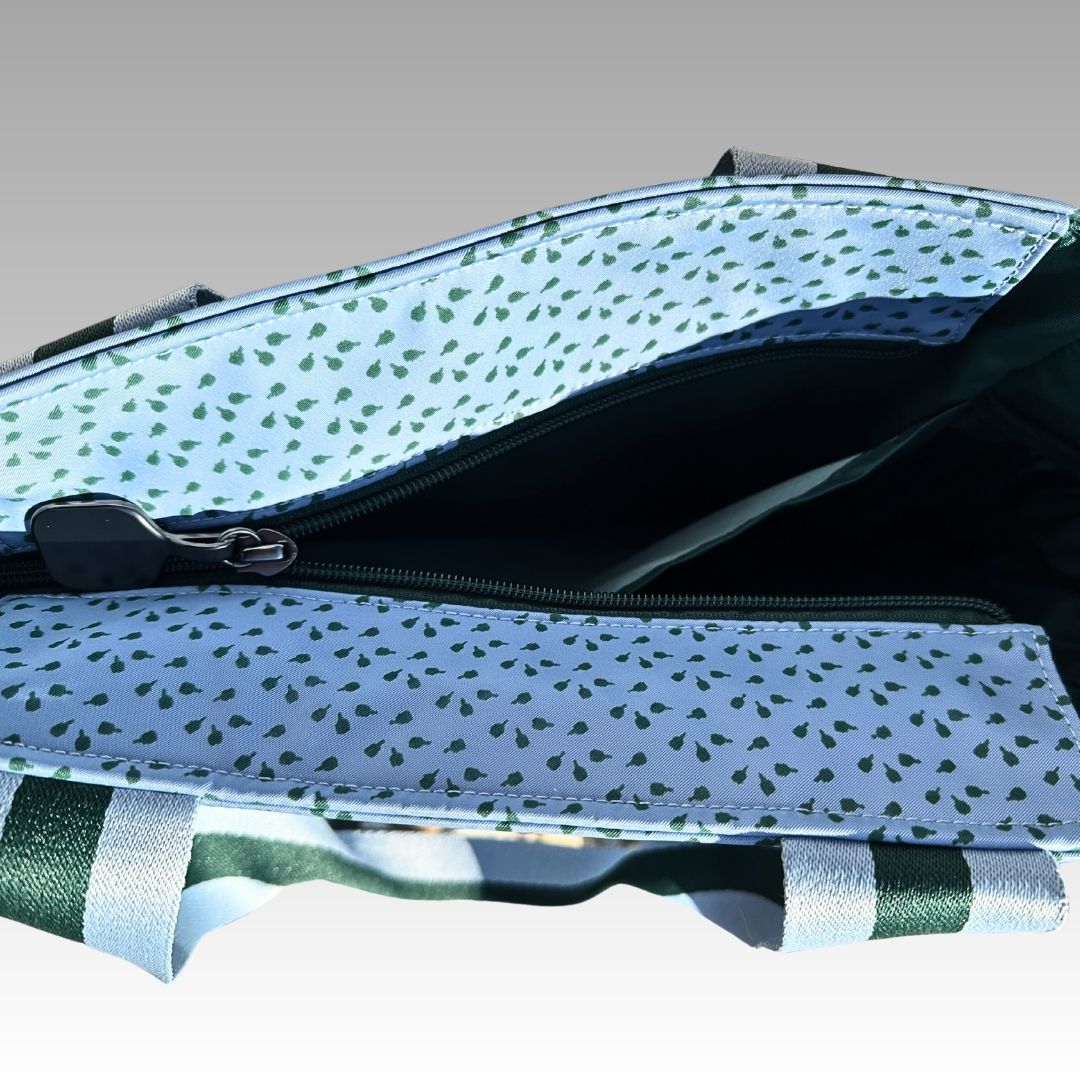 A close-up of the Playly Store’s Lulu Pickleball Tote shows its white interior with green leaf patterns, an inner zipper pocket, and sturdy green-and-white striped handles. The partially visible exterior appears against a gradient gray background.