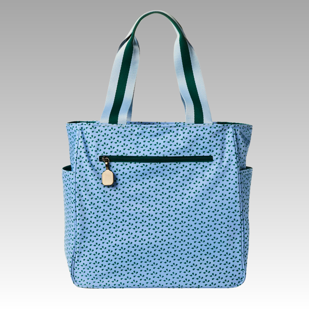 The Playly Store Lulu Pickleball Tote features light blue fabric with dark blue hearts, wide white handles with green stripes, a gold zippered front pocket, and two side pockets, displayed on a gradient gray background.
