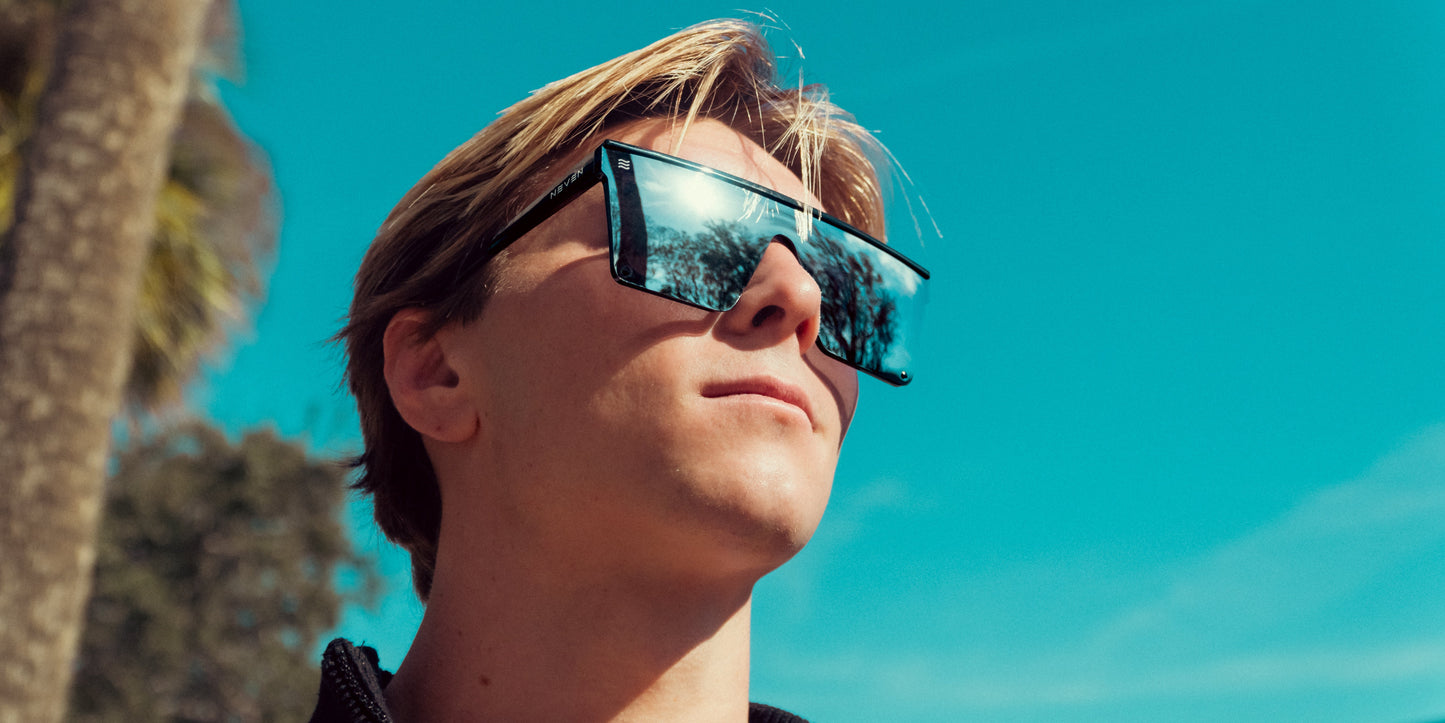 A young person with blond hair wears Neven Eyewear's Cr sunglasses, showcasing a modern shield design. The large, chromium frames reflect palm trees and a bright blue sky, hinting at a sunny outdoor setting.