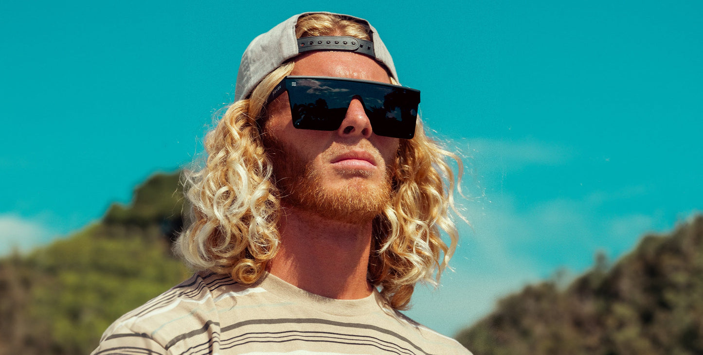 A man with long curly blond hair and a reddish beard wears a backwards gray cap, striped shirt, and Neven Eyewear IWMSAN polarized sunglasses (UV400 protection). He stands outdoors under a bright blue sky with blurred green hills behind.