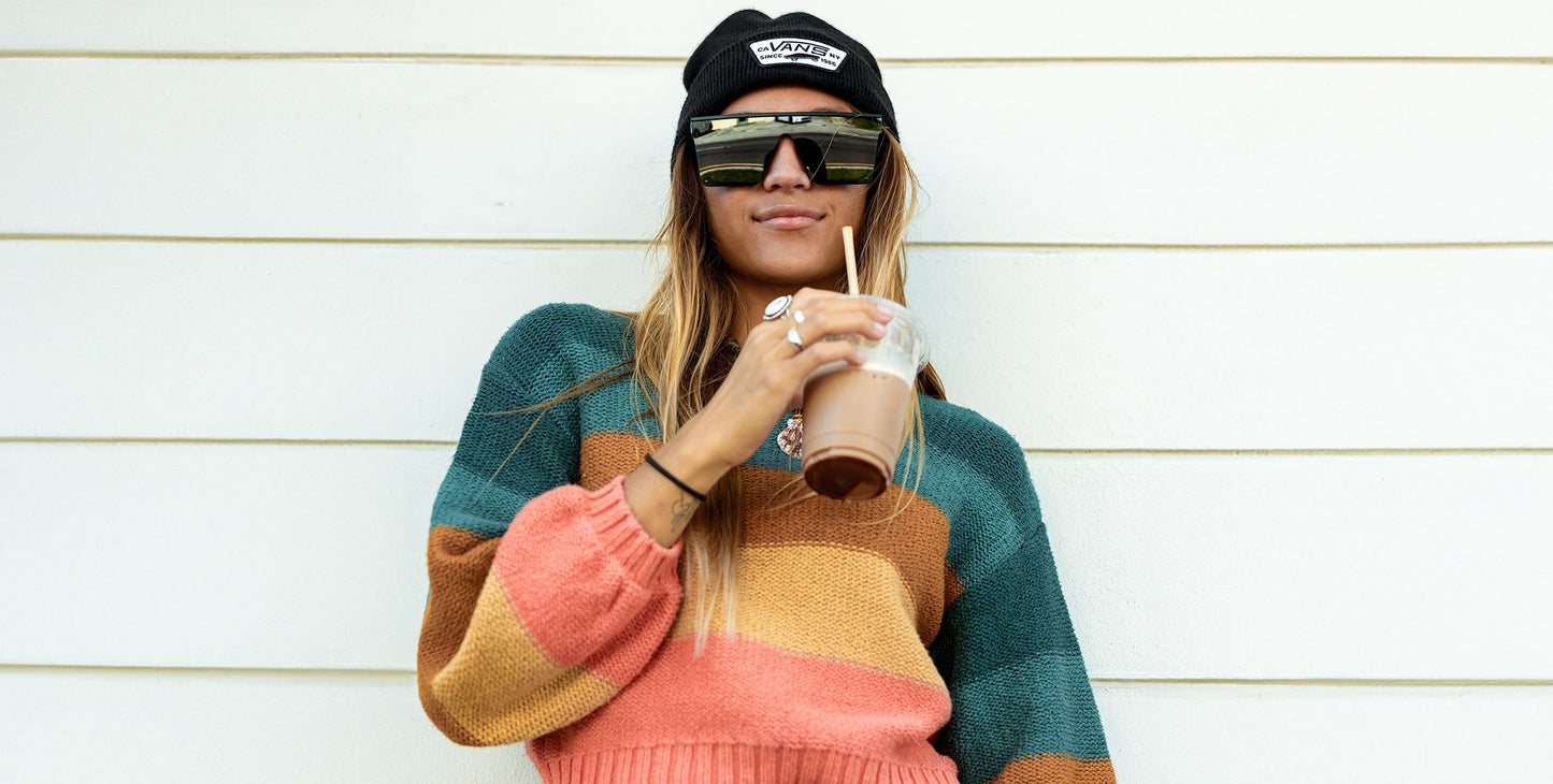 Wearing Neven Eyewear's IWMSAN sunglasses with UV400 protection, a woman in a striped sweater and black beanie stands against a white wall, holding iced coffee. She gazes down at the camera with long hair and a relaxed vibe.