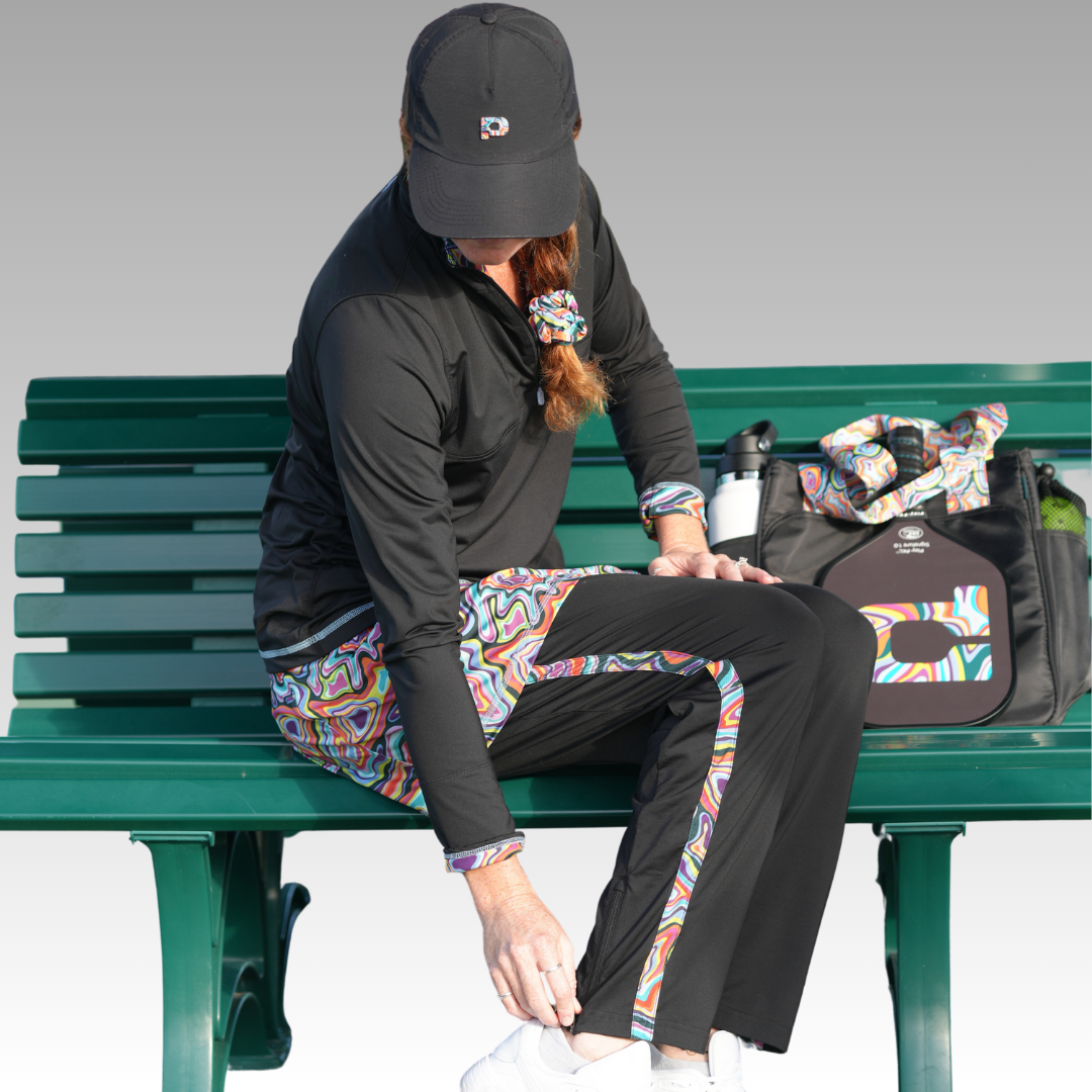 A woman in a black cap and colorful Playly Store activewear, including Lexi Warm-Up Pants with ankle zippers, sits on a green bench adjusting her white sneaker. Her auburn ponytail is tied with a matching scrunchie. A stylish bag rests nearby.