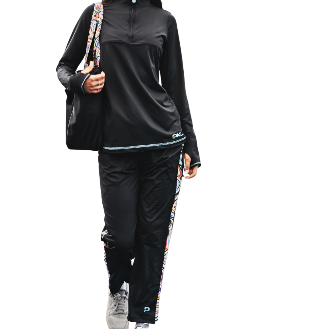 Wearing Playly Store's Lexi Warm-Up Pant with colorful side stripes and ankle zippers, a person in a black SwiftForm long-sleeve top and gray sneakers walks forward carrying a black shoulder bag, exuding casual, sporty style against a gradient gray background.