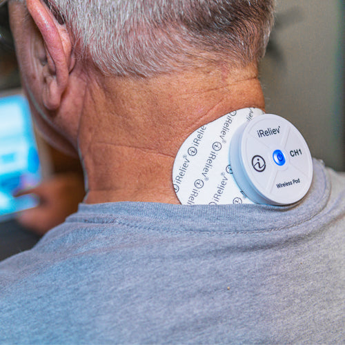 A person with short gray hair wears a gray shirt and uses the iReliev Wireless Large Single Pads Refill Kit, secured to their neck. The blue light indicates the device is active, set against a softly blurred background.