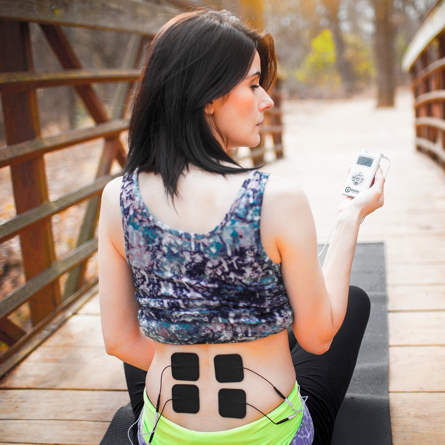 A dark-haired woman sits on a mat on a wooden bridge outdoors, facing away. She uses an iReliev Wired Large & Small Refill Kit, with four electrode pads on her lower back connected by wires for pain relief.