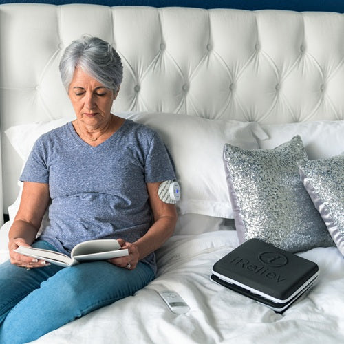 A woman with short gray hair reads on her bed in a bright, cozy room. Next to her are silver pillows and an iReliev Hard Protective Carry Case for Wireless TENS EMS, showcasing its practical use in a comfortable home setting.