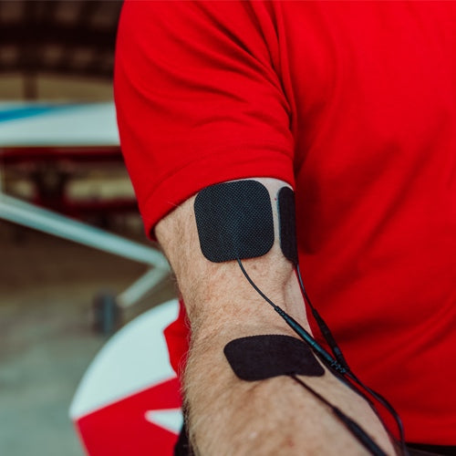 A person in a red shirt has three black iReliev TENS Unit Pain Relief System electrodes attached to their forearm and upper arm. In the background, part of a white airplane with red and blue markings is visible.