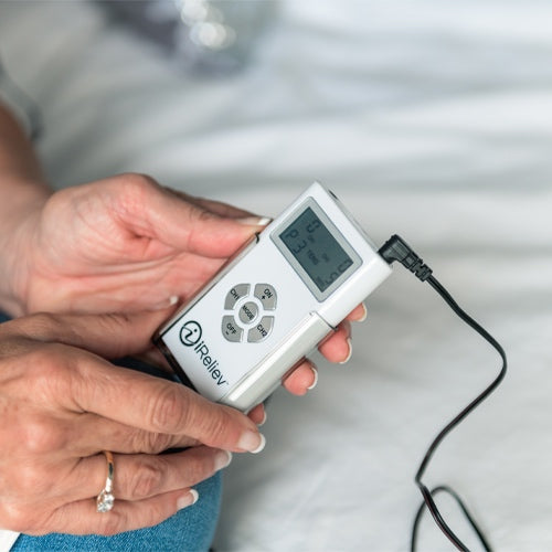 A person with manicured nails and a ring holds an iReliev TENS Unit Pain Relief System, featuring a small screen and control buttons, connected by a black wire—ready to use for pain relief while seated on a bed or couch.