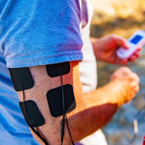 A person uses the iReliev Wired Large & Small Refill Kit, with four black electrode pads on their upper arm connected by wires to a handheld device, indicating electrical muscle stimulation therapy.