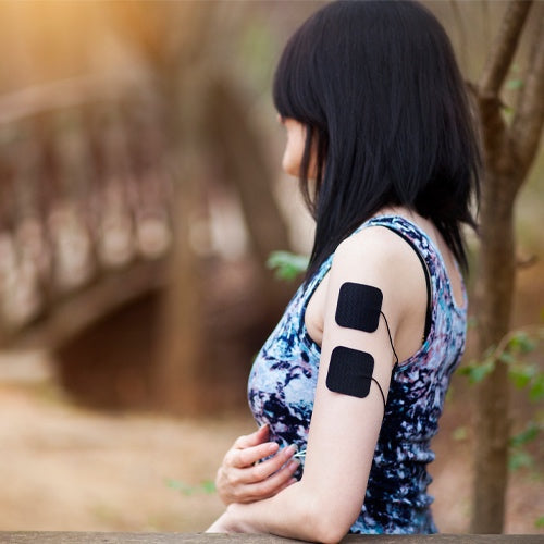A woman with straight dark hair, in a sleeveless patterned top, sits outdoors by a wooden bridge. She uses two black iReliev TENS + EMS Pain Relief & Recovery System pads on her upper arm, surrounded by blurred greenery.