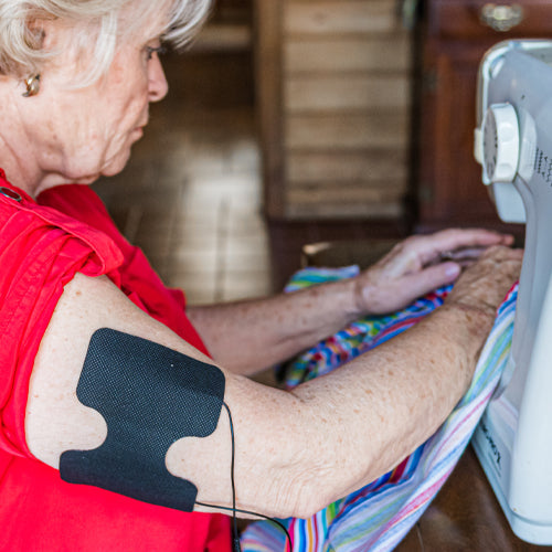 An older woman in a red shirt uses a sewing machine while an iReliev Wired Super Pads Refill Kit black electrode pad is attached to her upper arm with a wire. Colorful fabric is beneath the needle and the background is softly blurred.