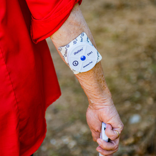 A person in a red shirt has an iReliev Wireless Large and Small Electrode Pads Refill Kit patch on their forearm. They hold a small device, possibly a remote, and are outdoors with grass and dirt in the background.
