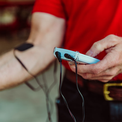 A person in a red shirt uses iReliev Extra Long TENS Lead Wires with pin connectors to attach electrodes to their bare forearm, indicating the use of a portable muscle stimulator. The white device and background appear blurry.