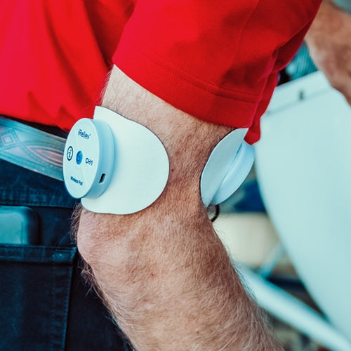 A close-up of a person’s forearm in a red shirt and patterned belt shows an iReliev Wireless Large Single Pads Refill Kit attached to the elbow, featuring buttons and indicator lights, secured by multi-layer adhesive patches.