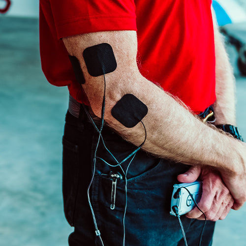 A person wearing a red shirt uses the iReliev Wired Small Pads Refill Kit, with black patches and wires attached to their arms, holding an electronic device—likely for muscle stimulation or physical therapy.