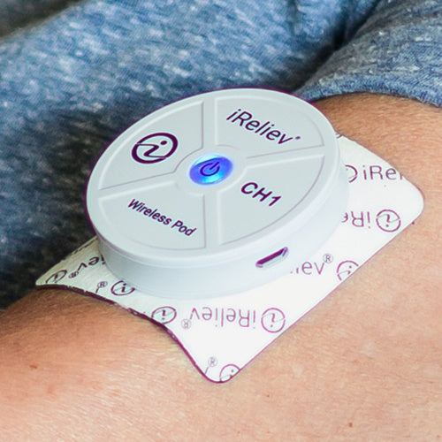 A round, white iReliev Wireless Small Pads Refill Kit pod with a blue power button and logo is attached to a person’s skin using a multi-layer adhesive pad. The person is wearing a grey fabric shirt.