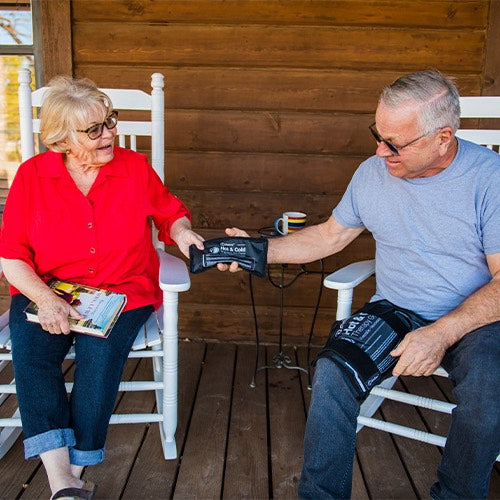 An older woman in a red shirt and glasses smiles at an older man checking her blood pressure as they sit on the porch. They discuss the iReliev Premium Reusable Hot and Cold Therapy Gel Pack with Securement Straps for pain relief.
