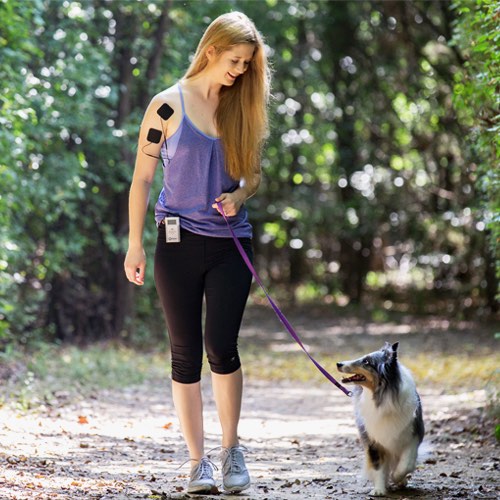 A woman with long blond hair in a purple tank top walks her black-and-white dog on a trail, wearing two black electrode patches on her upper arm connected to the iReliev TENS + EMS Pain Relief & Recovery System clipped to her waistband.
