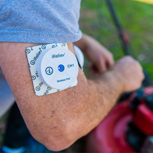 Wearing a gray shirt outdoors, a person uses an iReliev Wireless Small Pads Refill Kit on their upper arm while mowing the lawn, with grass and blurred greenery in the background.