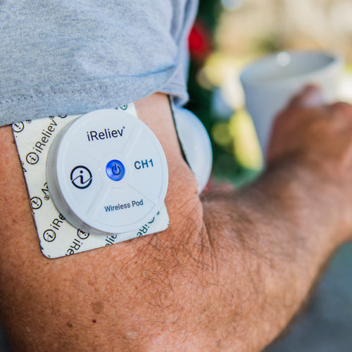 A close-up shows a person’s upper arm with an iReliev Wireless Large and Small Electrode Pad from the Refill Kit attached to their skin. The pod is labeled “CH1,” has a power button, and they hold a cup in the other hand; background is blurred.