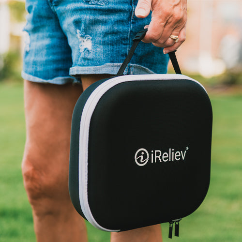 A person in ripped denim shorts holds a black iReliev zippered case, ideal for storing the iReliev Percussion Massage Gun, against a grassy outdoor background.