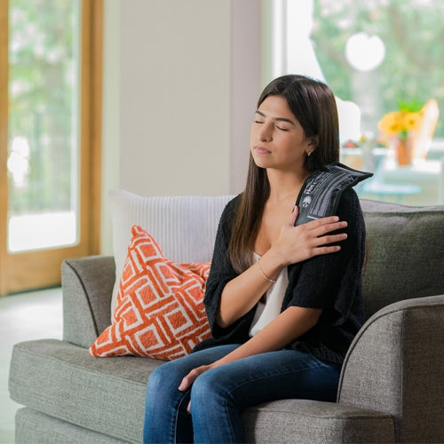 A young woman with long dark hair sits on a gray couch in a bright room, using the iReliev Hot and Cold Bundle on her left shoulder for pain relief. She wears a black sweater and jeans; an orange-patterned pillow is nearby.