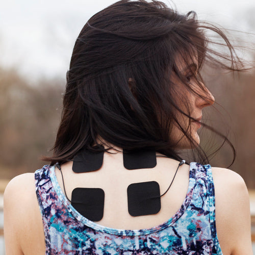 A woman with dark hair wears a sleeveless, patterned top outdoors. She uses iReliev Wired Small Pads Refill Kit, with four black electrode pads and wires attached to her neck and upper back.