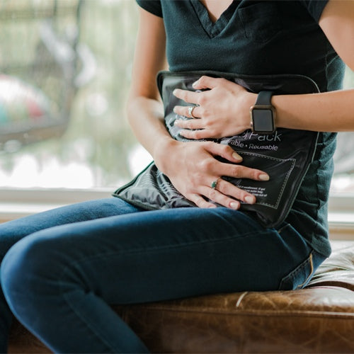 Indoors, a person in a black t-shirt and jeans sits on a brown cushion, wearing rings and a smartwatch, holding the iReliev Premium Reusable Hot and Cold Therapy Gel Pack with Securement Straps against their lower abdomen for pain relief.