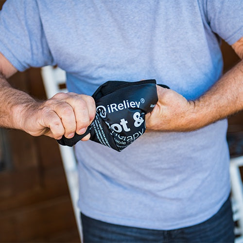 A person in a light gray t-shirt wrings a black iReliev Premium Reusable Hot and Cold Therapy Gel Pack with Securement Straps. The background is blurred, showing a wooden surface and a white chair.