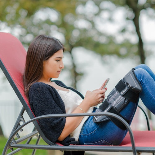 A woman in casual wear relaxes on a red outdoor lounge chair, using her phone. She has the iReliev Premium Reusable Hot and Cold Therapy Gel Pack with Securement Straps wrapped around her right knee for pain relief.