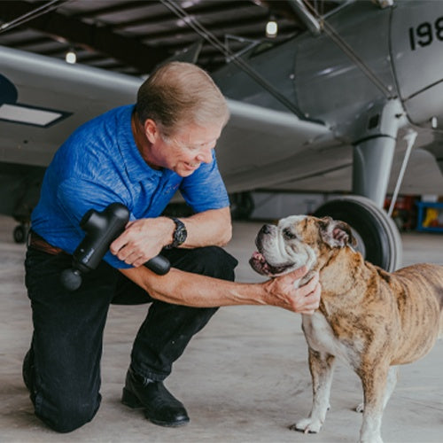 A smiling man in a blue shirt kneels beside a happy bulldog, gently holding its chin. In his other hand, he holds the iReliev Percussion Massage Gun. They are inside a hangar with a vintage gray airplane in the background.
