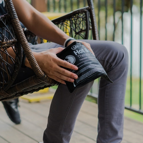 A person sits on a wooden porch swing holding the iReliev Hot and Cold Bundle against their knee for pain relief. They wear gray pants and a black watch, with greenery and a metal railing blurred in the background.