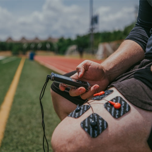 A person sits on a sports field using the iReliev SPORT Muscle Stimulator with Explosive Strength Mode, attached to their thigh with four black electrode pads and red wires, holding the controller. A running track and green grass are visible in the background.