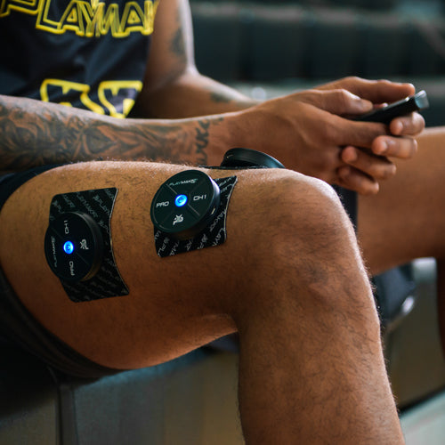 A close-up of a tattooed thigh shows two iReliev PRO Wireless Muscle Stimulators with TENS + EMS, blue lights on, attached. The person in a black and yellow outfit holds a remote, focusing on muscle recovery.