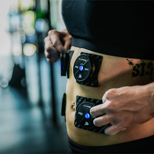 A close-up shows a person adjusting two black iReliev PRO Wireless Muscle Stimulator with TENS + EMS devices, their blue lights visible on the abdomen, suggesting muscle recovery use in a blurred fitness setting.