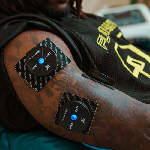 A person’s arm with two square black iReliev PRO Wireless Muscle Stimulator with TENS + EMS pads attached to the bicep and forearm. The pads have blue LED lights, white text, and the person wears a black mesh sports jersey with yellow writing.