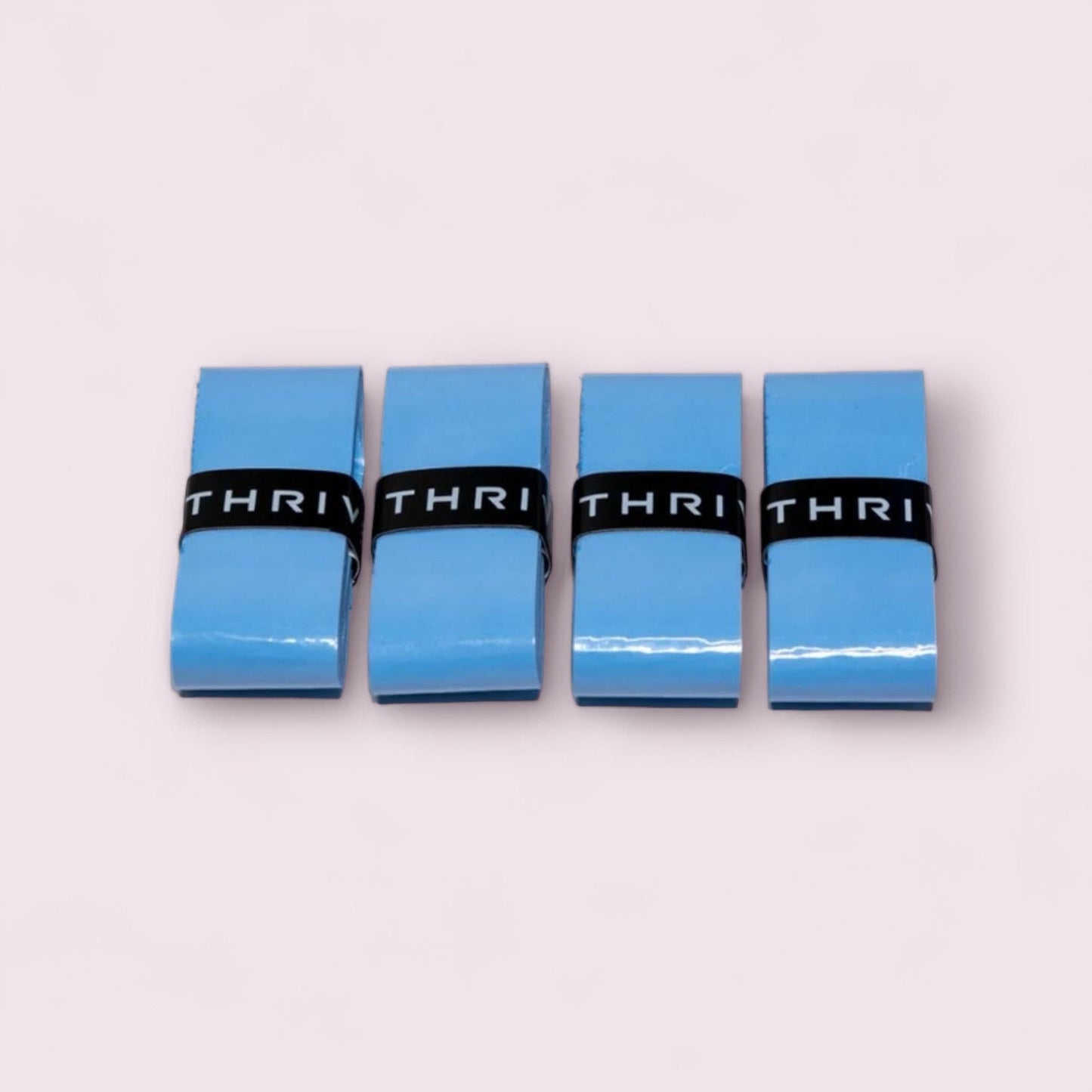 Four neatly folded blue Super Tacky Overgrips from Thrive Pickleball are arranged side by side on a light pink background. Each durable overgrip is wrapped with a black strap labeled THRIV in white capital letters.