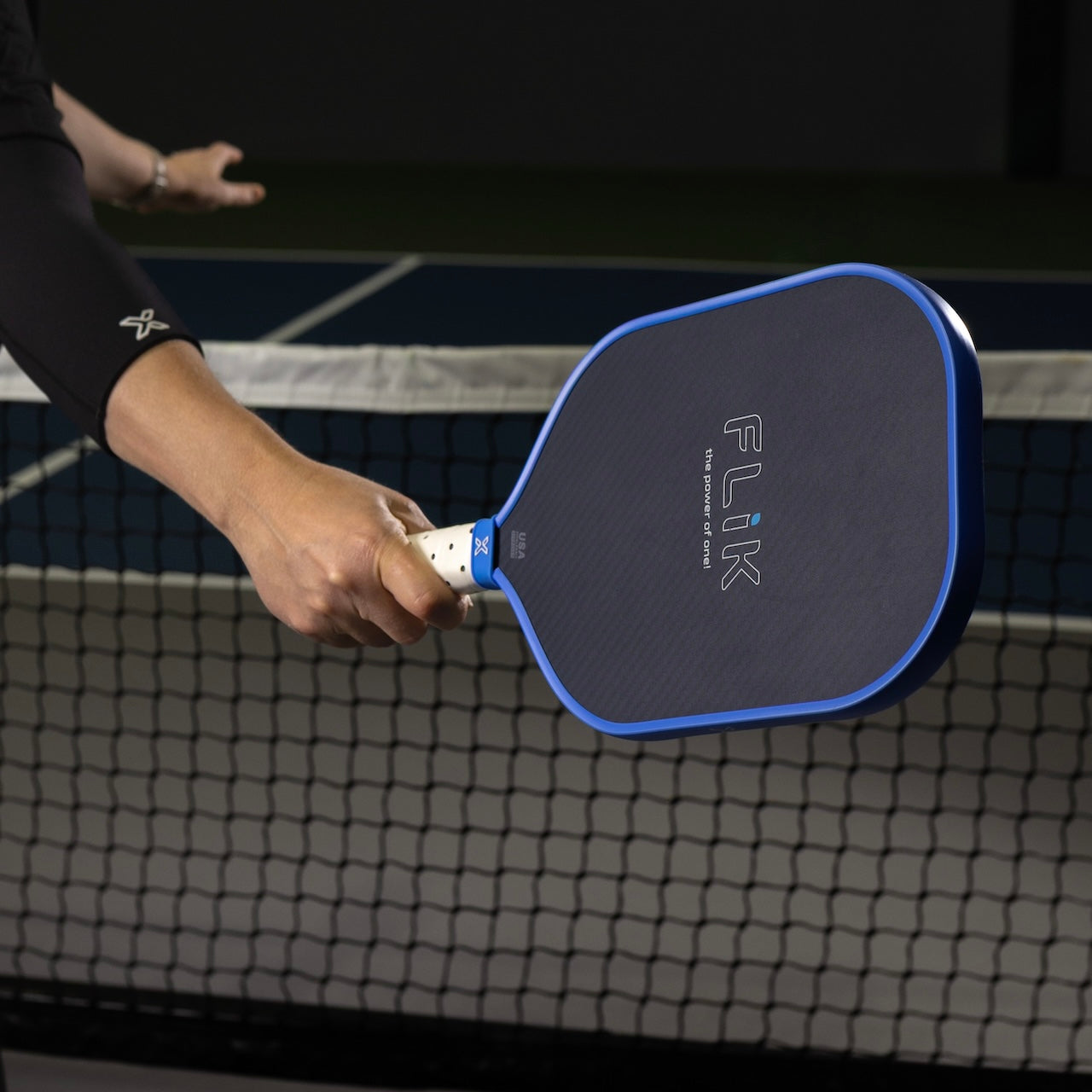 A hand grips the body helix FLIK Gen 3 Pickleball Paddle—USAP Approved, featuring a blue edge and cushioned grip—poised over an indoor net, ready to hit or just after hitting a ball in the dimly lit court background.