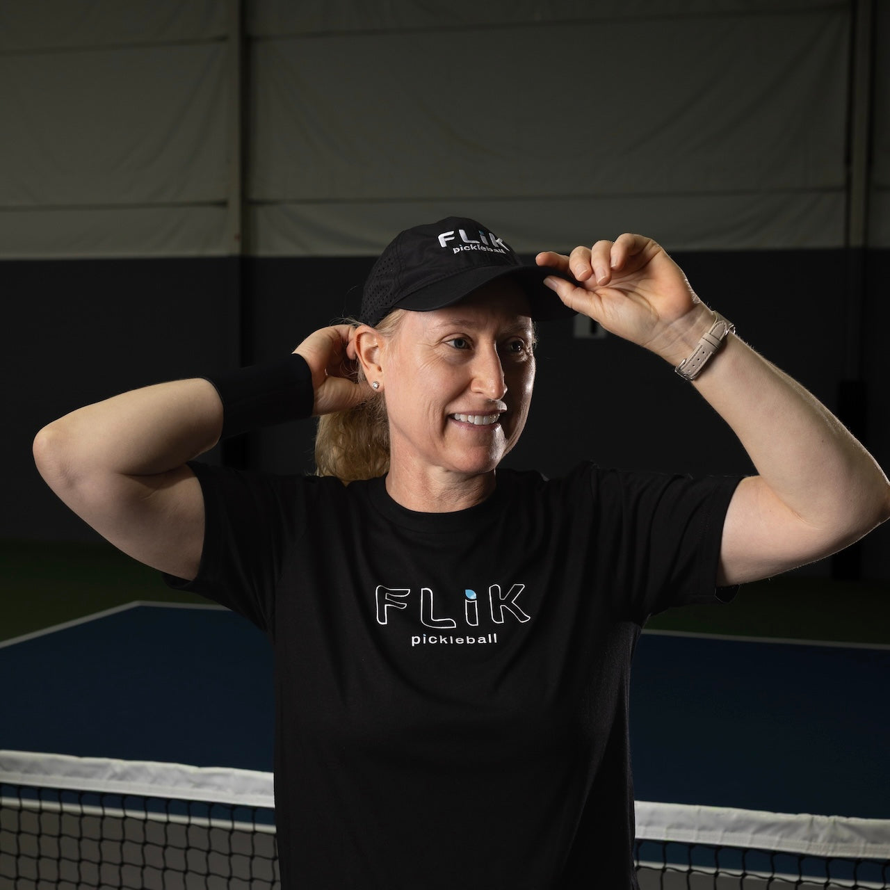 On an indoor court, a smiling person adjusts a black body helix FLiK Pickleball Hat, pairing it with a matching logo tee, black wristband, and smartwatch. The net and dark walls set the background scene.