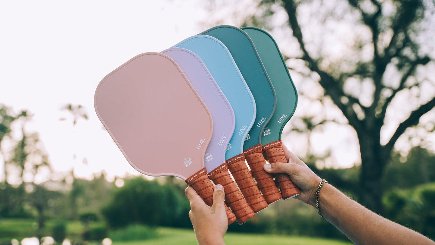 A person holds up five matte pastel LUXE Pickleball Core Collection carbon fiber paddles with brown leather-wrapped handles outdoors. The textured surfaces offer better grip and control. Blurred trees and greenery are seen in the background.