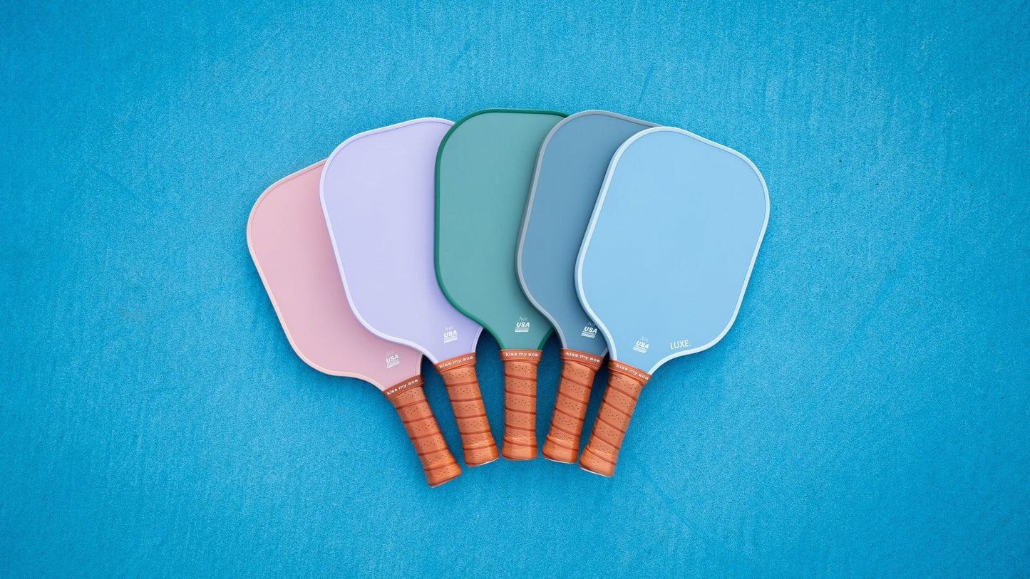 Five LUXE Pickleball Core Collection paddles with brown handles are fanned out on a blue surface, each in solid pastel—pink, lavender, green, teal, and blue—with a textured face for better grip and control. USA Pickleball Approved.