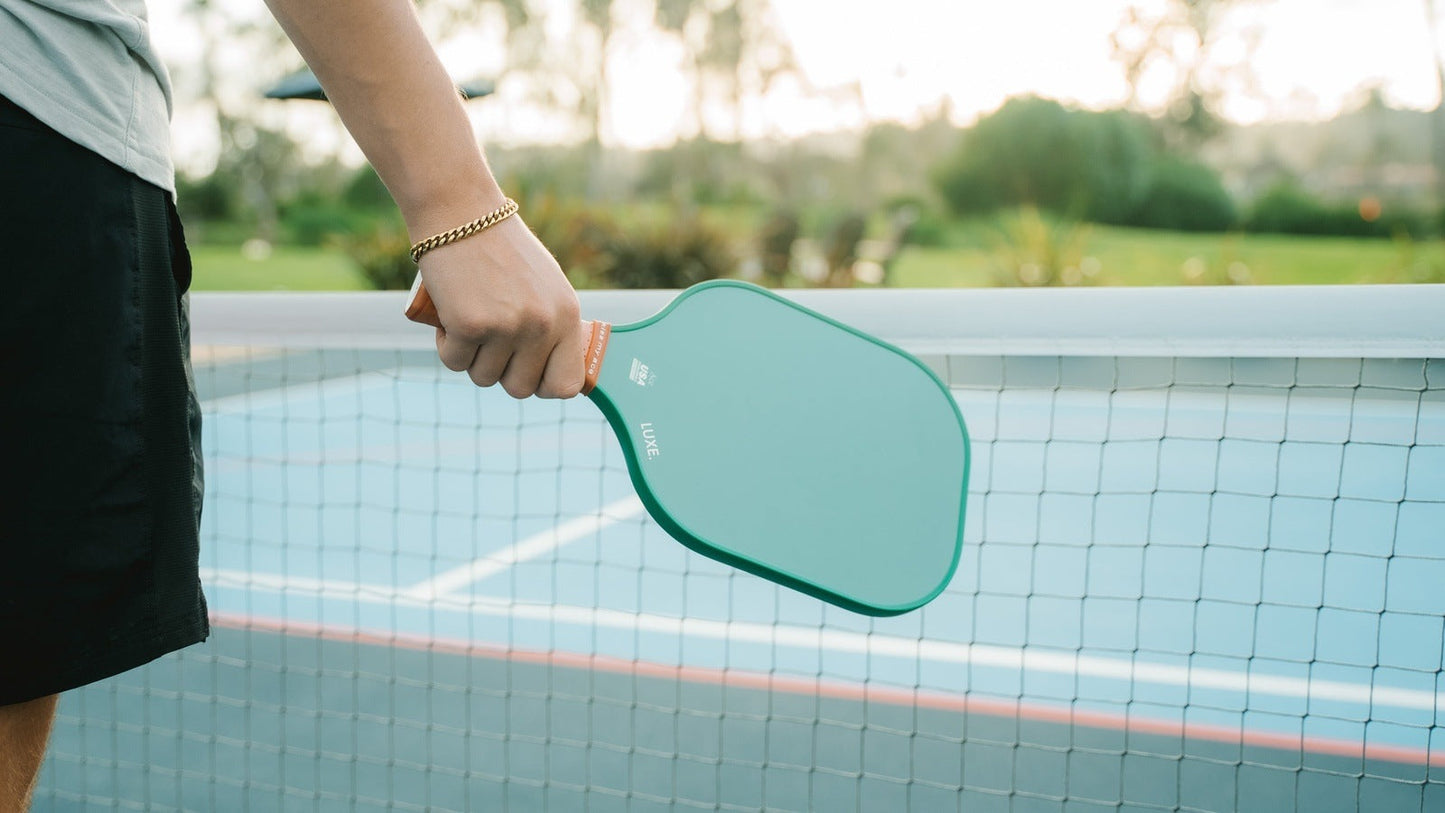 A person in black shorts and a gold bracelet stands by a net on an outdoor court, holding the LUXE Pickleball Core Collection paddle with its textured green carbon fiber surface. Soft sunlight highlights greenery and blurred trees in the background.