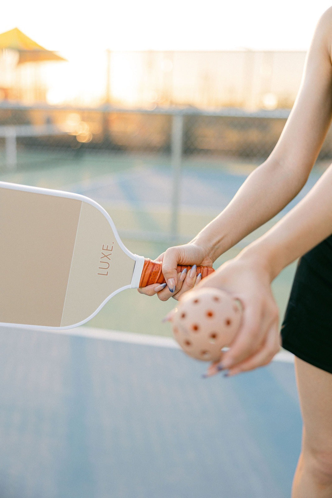 A person holds a beige LUXE Pickleball Dipped - Ace paddle in one hand and an orange perforated ball in the other, ready to serve on an outdoor court with soft sunlight and stylishly blurred lines, creating an inviting scene.
