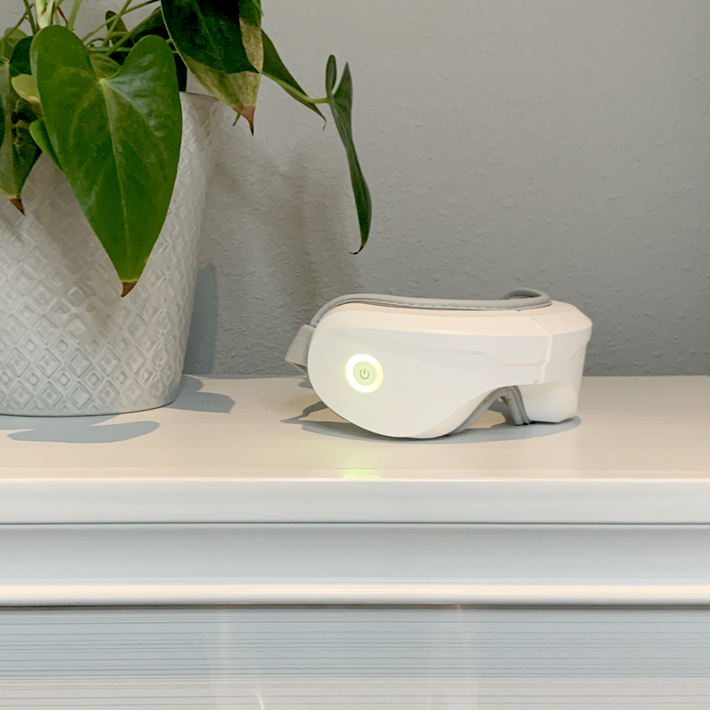 The iReliev Relaxable™ Eye Massager with Heat, featuring a glowing green power button, rests on a glossy white dresser beside a potted plant with large green leaves; the scene is set against a plain light gray wall.