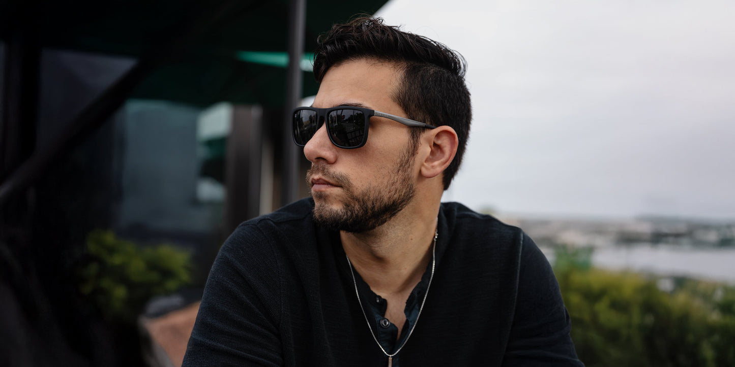 A man with dark hair and a beard wears Neven Eyewear's Blueprint black polarized sunglasses with UV protection, a dark shirt, and a necklace. He sits outdoors, looking left, greenery and a modern building softly blurred behind him.