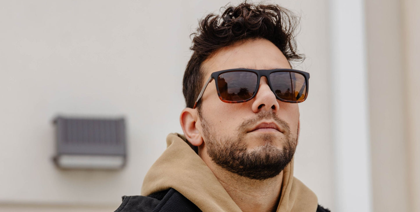 A man with short, dark, curly hair and a beard wears Neven Eyewear Repeater sunglasses with polarized lenses and a tan hoodie under a dark jacket, standing outdoors by a light wall with a gray outdoor light fixture behind him.