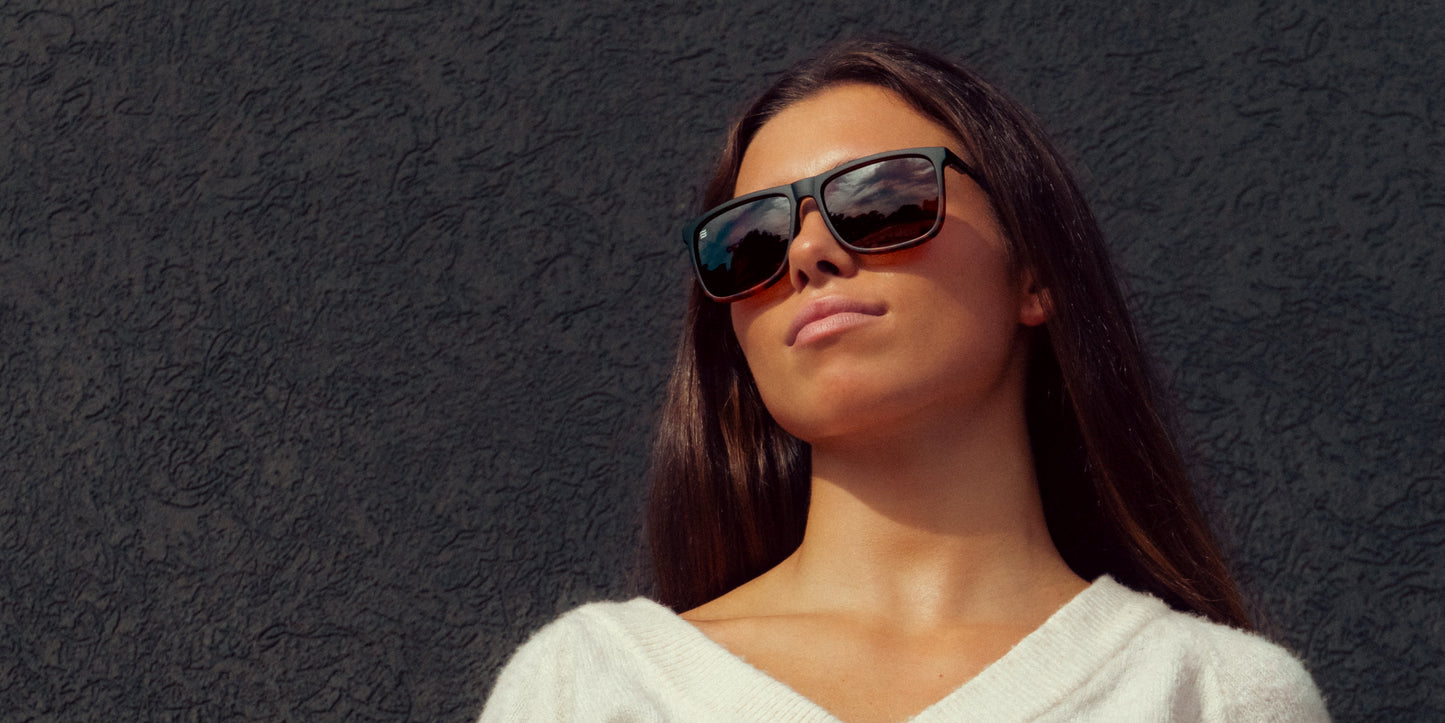 A woman with long dark hair wears large black polarized Repeater sunglasses from Neven Eyewear and a white V-neck sweater, standing against a textured black wall as cloud reflections shine in her shades while she gazes upward confidently.
